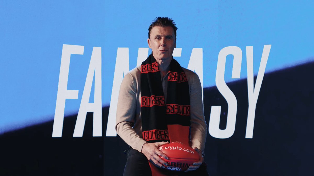 Matthew Lloyd, wearing an Essendon Bombers scarf and holding a Crypto.com-sponsored football, stands in front of a blue backdrop displaying "FANTASY" in large white letters, aligning with the post's promotion of an AFL Grand Final fantasy contest.
