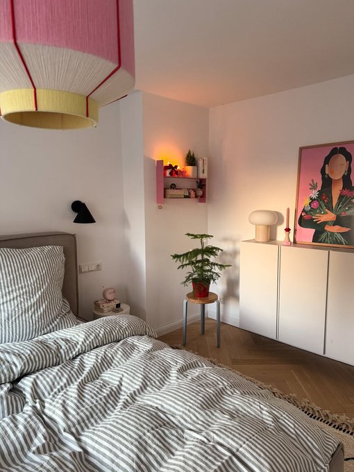 A cozy bedroom with a gray striped bed, a small Christmas tree, white cabinets, a pink shelf, and a framed pink-toned portrait of a woman on the wall.
