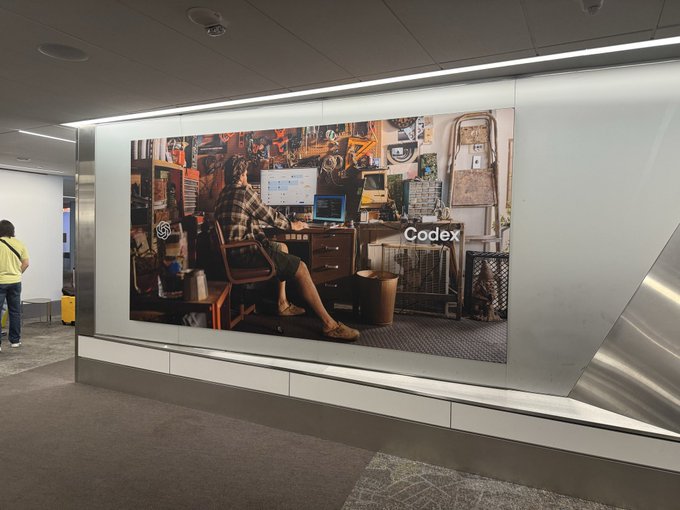 A large illuminated advertisement displays the word 'Codex' alongside an image of a person working at a cluttered desk with multiple computer monitors, mounted on a wall in what appears to be a public space.