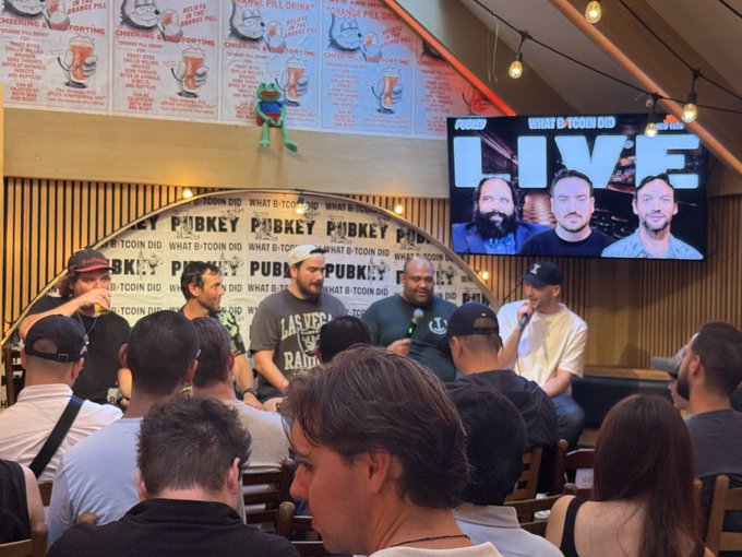 A live recording of "What Bitcoin Did" podcast at PubKey, featuring several panelists seated on stage with microphones, while a large screen displays the show's "LIVE" branding alongside headshots of the hosts, and the audience watches from the venue. The backdrop is covered with beer-related posters and PubKey branding, matching the live event context described in the post.
