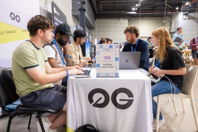 Several people sit at a table covered with a white '0G' tablecloth, working on laptops and engaging in conversation at what appears to be a tech conference.