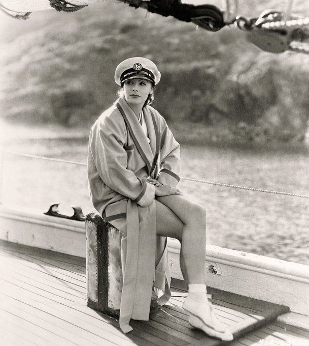 A vintage black-and-white photograph of a woman in a sailor cap and draped shawl sitting on the deck of a wooden boat by the water.