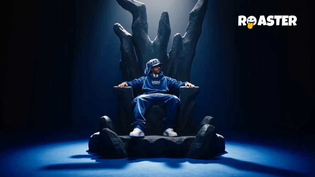 The image shows a man sitting in a chair with his feet off of it. The chair appears to be an oversized or throne-like seating arrangement, adding an air of importance or significance to the scene. The man's attire includes a blue jacket and pants, and he is seated comfortably in front of what looks like a TV screen. In the background, there are two billboards featuring images related to the post: one with a man wearing a crown, symbolizing the title "Seeker", and another with an image of a man on a throne, representing the title "The duopoly" or "the gatekeepers". The overall setting suggests that this could be part of a promotional campaign for the movie "Seeker", which is known for its gripping drama and intense conflicts.