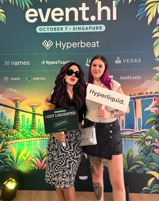 Two women posing at the 'event.hl OCTOBER 7 · SINGAPURR' crypto event, holding signs that read 'I GOT LIQUIDATED [AGAIN]' and 'Hyperliquid.' in front of a sponsor backdrop featuring Hyperbeat, VEGAS, and other logos.