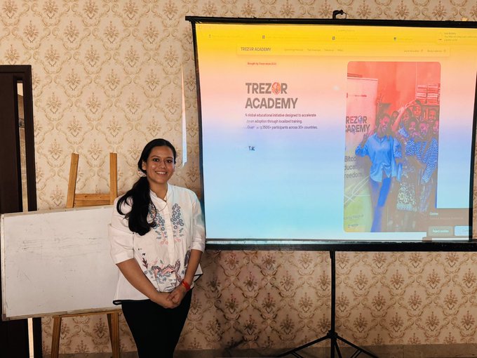 A woman in a white embroidered blouse stands smiling next to a projection screen displaying the 'TREZOR ACADEMY' website for an educational initiative on blockchain and Bitcoin self-custody.