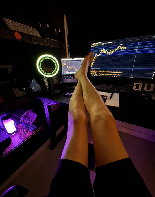 The image shows a person's feet propped up on a desk with their legs crossed while sitting at a computer. The desk also has a laptop and a monitor displaying stock market data. The person appears relaxed, possibly taking a break or working from home. The photo is related to the post by Northern Bro, which explains that many newbies fall into the misconception of chasing positive news about the funding rate for a company when it's actually a reflection of the overall bullish sentiment in the market. This can lead to poor investment decisions and financial losses if not properly understood.