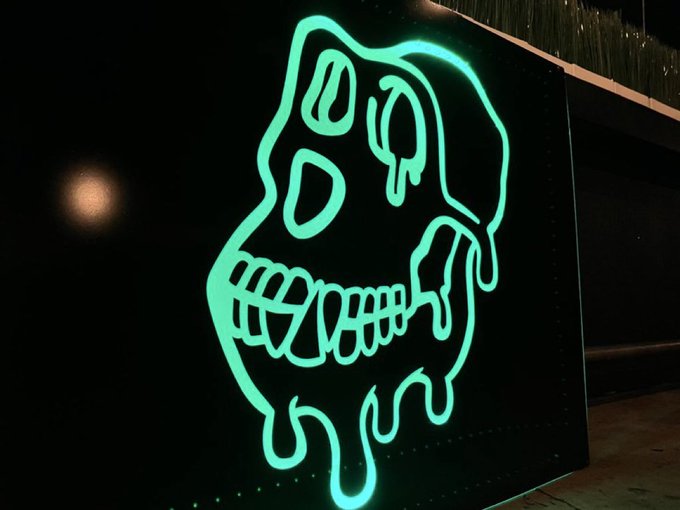 A neon green light sculpture depicting a melting skull with the word 'DOOM' incorporated into the teeth design, mounted on a dark panel at night.