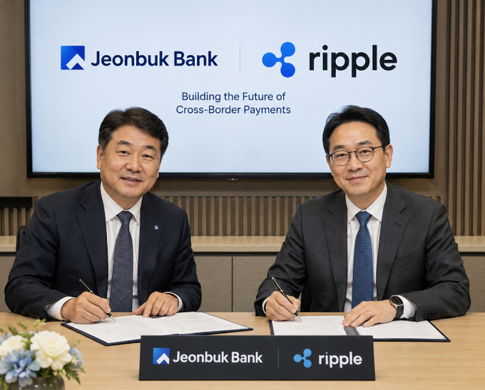 Two men in suits sitting at a table signing documents, with a screen behind them displaying logos for 'Jeonbuk Bank' and 'ripple' alongside the text 'Building the Future of Cross-Border Payments'.