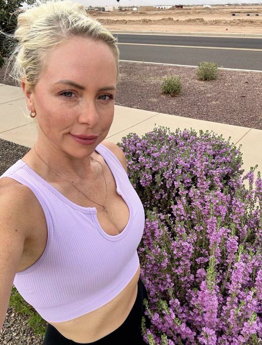 A selfie of a blonde woman in a light purple sports top posing next to a blooming purple flowering bush outdoors.