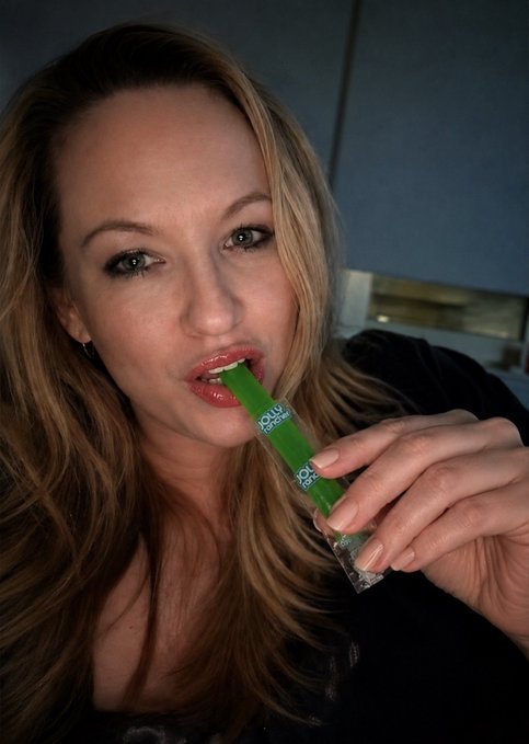 A close-up photo of a woman with blonde hair holding a green popsicle-like product near her mouth.
