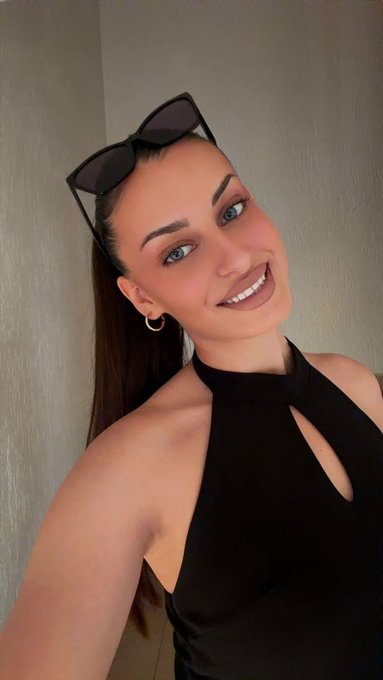 A young woman with long brown hair, wearing a black halter top with sunglasses perched on her head, smiles warmly in a selfie.