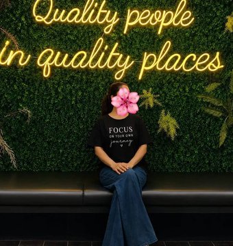 A person with a pink flower emoji covering their face, wearing a black 'FOCUS ON YOUR OWN journey' t-shirt and jeans, sitting in front of a green hedge wall with neon text reading 'Quality people in quality places'.