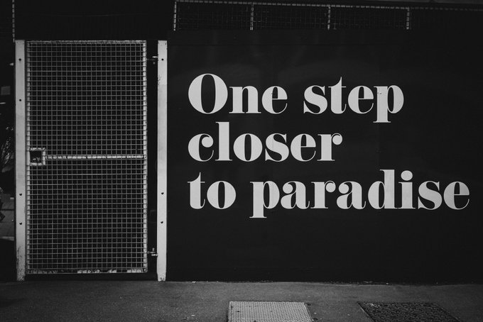 A black wall with white text reading 'One step closer to paradise' next to a metal mesh gate.