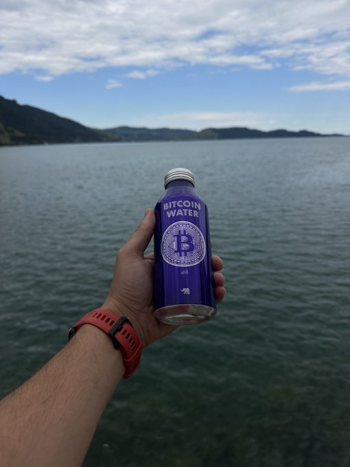 A hand holds a can of "Bitcoin Water" with the Bitcoin logo, emphasizing the "Bitcoin Forever" sentiment from the accompanying post. The serene water and landscape provide a backdrop to this crypto-themed beverage.