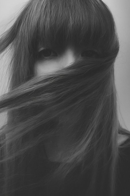 A black-and-white moody portrait of a person with long bangs covering the upper half of their face, while windblown hair sweeps across and obscures the lower portion of their face, leaving only their eyes visible.