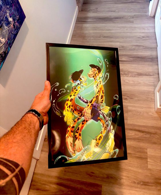 A tattooed arm holds up a framed, colorful illustration depicting two leopard-print humanoid figures in a dynamic embrace against a green background, displayed on a wooden floor.