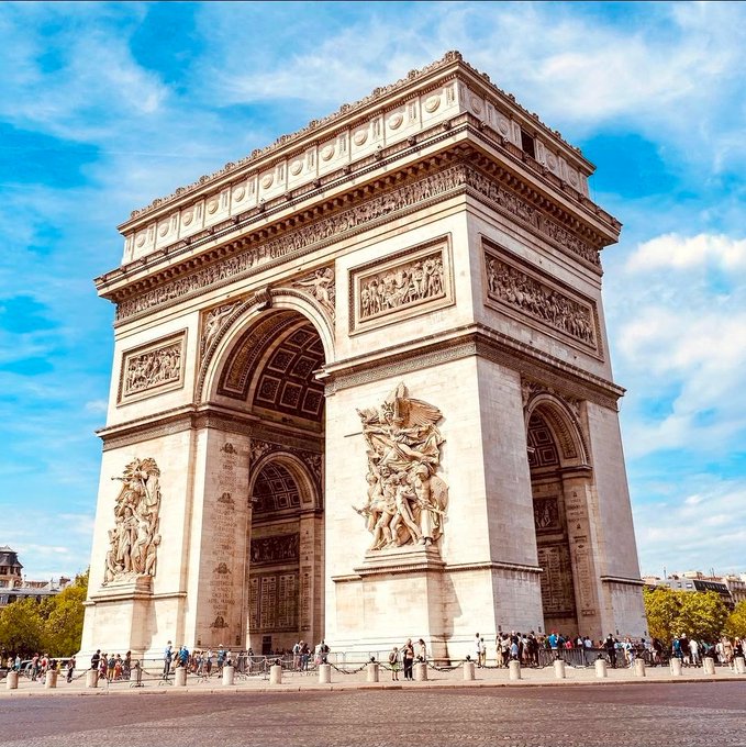 The image features a large white archway with intricate carvings of people and animals, standing tall against a blue sky with clouds. The archway is part of a larger complex that includes other buildings nearby. There are several people walking around in front of the archway, possibly admiring its design or going about their day. A few individuals can be seen carrying handbags as they explore the area.