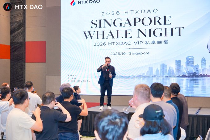 A man speaks on stage at an 'HTX DAO' event, holding a microphone in front of a screen displaying '2026 HTXDAO SINGAPORE WHALE NIGHT' and '2026.10 Singapore', with an audience seated in front.