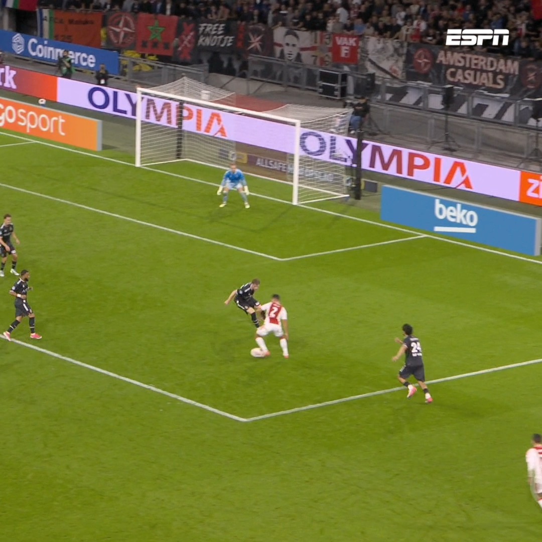The image shows an Ajax player in a white and red kit (number 2) controlling the ball near the goal area during a match, with the goalkeeper in blue and defenders in dark kits positioned around the box, and visible advertising boards reading "OLYMPIA" and "beko." This action shot captures the moment of a scoring opportunity that aligns with the post's celebration of Julian Brandt's goal-scoring form for Ajax, showing him in a dangerous attacking position close to goal.