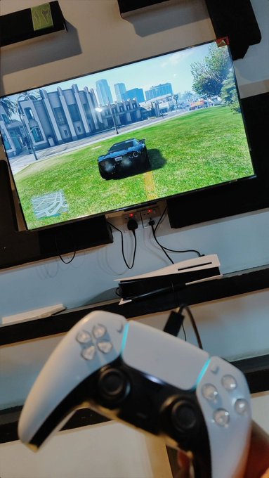 A first-person view of someone holding a white PlayStation 5 DualSense controller in front of a TV displaying a video game featuring a black sports car on a grassy field.