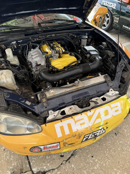 Open engine bay of a heavily branded Mazda race car, featuring a yellow engine cover, large black intake tube, radiator, and exposed wiring. The front bumper displays large “mazda” lettering along with “FALKEN” and “PIAA” stickers, while a “mazda Raceway Laguna Seca” sign is visible in the background.