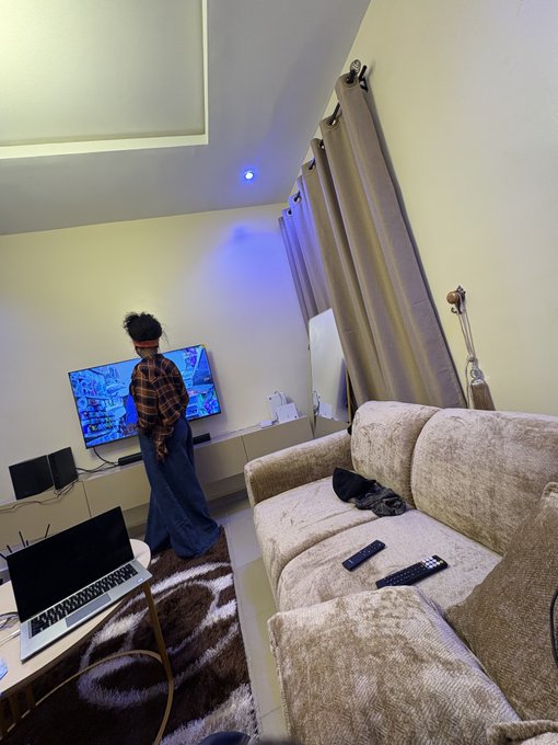 A young person stands in a modern living room watching a brightly lit TV displaying multiple colorful screens, while a laptop, remote controls, and a beige couch fill the foreground. The scene captures a casual mentoring visit, where the mentee is enjoying screen time in the cozy, tech-filled space.