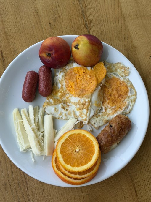 A white plate of food containing fried eggs, peaches, sausages, shredded cheese, orange slices, and a pastry, served on a wooden table.