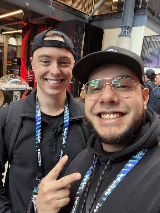 Two smiling young men pose for a selfie at what appears to be a convention, with one wearing a black backwards cap and the other wearing aviator glasses, both sporting lanyards with visible text.