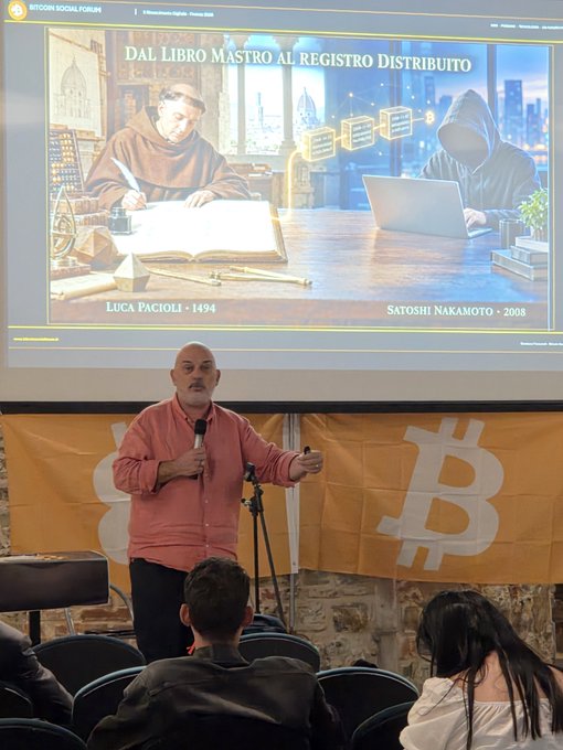 A bald man in a red shirt speaks at the Bitcoin Social Forum, with a slide behind him titled "DAL LIBRO MASTRO AL REGISTRO DISTRIBUITO" comparing Luca Pacioli (1494) to Satoshi Nakamoto (2008)—illustrating the historical evolution from double-entry bookkeeping to triple-entry distributed ledger systems like Bitcoin. The orange backdrop displays the Bitcoin "B" logo, tying the lecture's theme of accounting innovation to the cryptocurrency movement.
