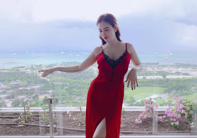 A woman in a red dress poses on a balcony overlooking a scenic ocean view with boats and greenery, complementing the post's "stay red" message—her vibrant red attire symbolizing the bullish sentiment expressed in the caption.