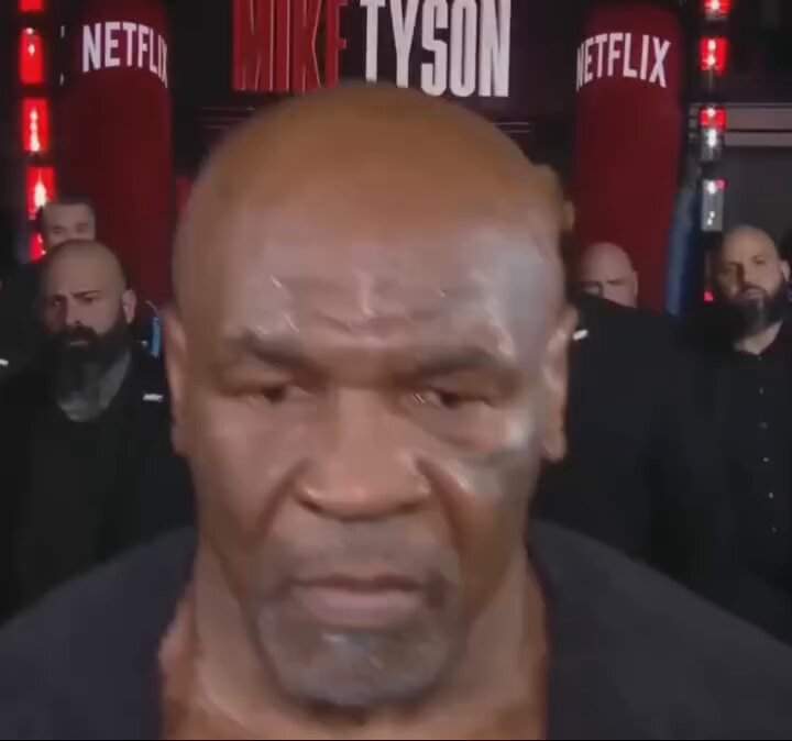 The image features a man with a shaved head, wearing a black shirt, standing in front of a boxing ring. The man appears to be a professional boxer or wrestler, possibly at a boxing event like the one shown on the screen. In the background, there are two men who seem to be part of the event as well. One of them is holding a microphone and talking into it while the other person is standing next to him.