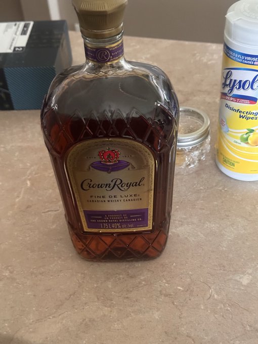 A photograph of a bottle of Crown Royal Fine De Luxe Canadian Whiskey sitting on a speckled countertop next to a container of Lysol Disinfecting Wipes and a small jar.