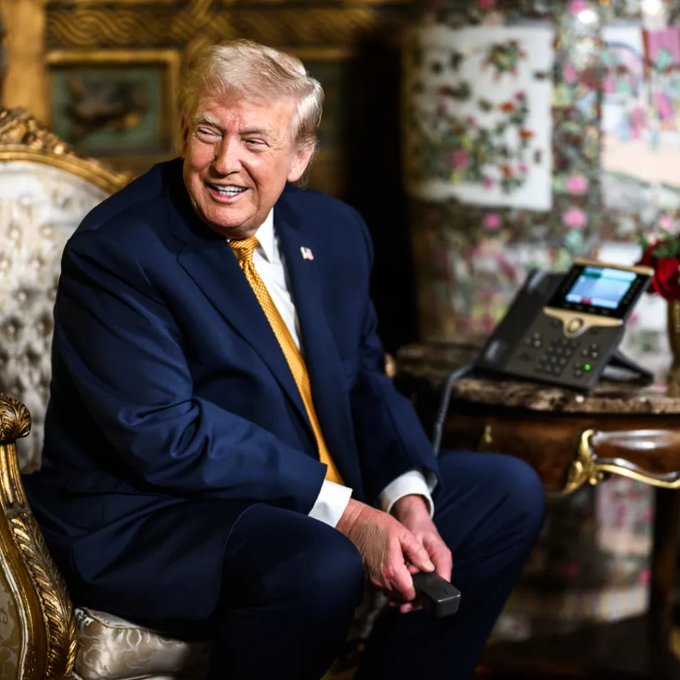 Donald Trump smiles while sitting in a chair, holding a remote, as he discusses his involvement in crypto "a little bit for politics." The image is a candid shot hinting at his recent comments on Bitcoin.