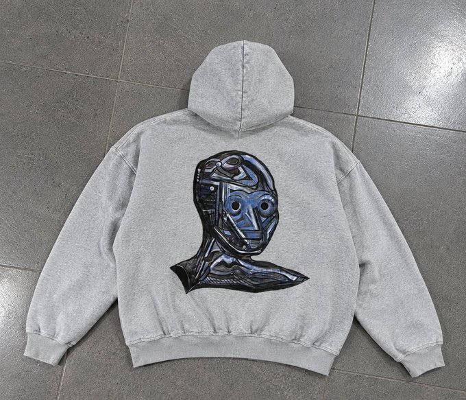 A light gray hooded sweatshirt lies flat on a tiled floor, featuring a large graphic of a robotic blue and black head on the back.