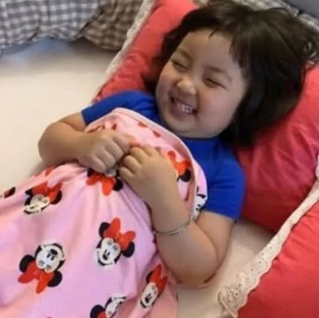 A young child with short dark hair, wearing a blue shirt and a pink Minnie Mouse blanket, smiling joyfully while lying on a bed with red pillows.