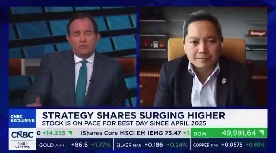 The image shows two men sitting behind a desk with their hands on their laps while they are talking about strategy shares surging higher. The first man has his arms crossed over his chest, and the second man is wearing a suit. They appear to be discussing business strategies or news related to Bitcoin.