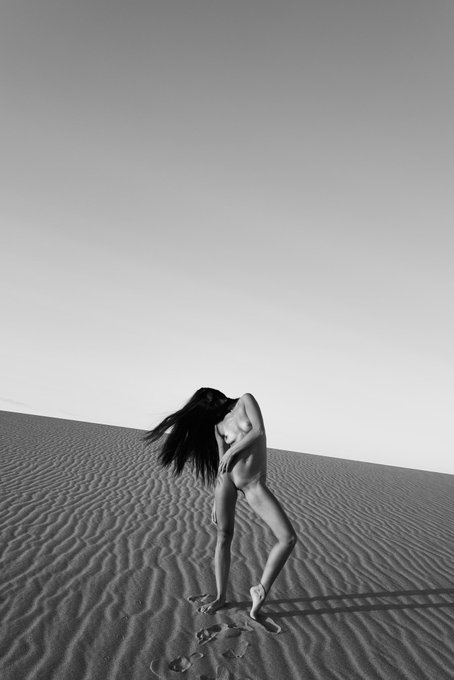 In this striking black and white photograph, a nude figure with long, flowing hair stands in a vast desert landscape, their form echoing the artistic journey of the photographer. The image serves as a visual representation of the artist's shift from NFT promotions to a personal visual diary.