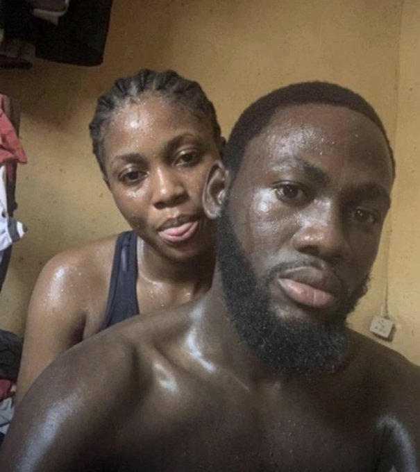 A selfie of two dark-skinned individuals, a man in the foreground and a woman behind him, both appearing to be sweating in a dimly lit room.