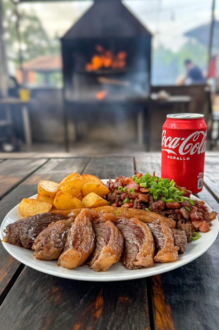 A plate of food featuring sliced grilled steak, roasted potatoes, and seasoned meat topped with green onions, served with a red can of Coca-Cola on a wooden table with a smoker/grill visible in the background.