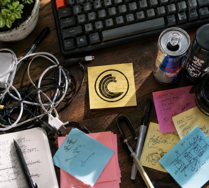 A cluttered wooden desk features a keyboard, tangled white earbuds, a Red Bull can, pens, and various colorful sticky notes including one that reads 'LAUNCH V2 - landing page - fix nav - icons' and another with a drawn 'C' logo.