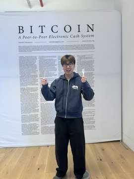 A person wearing a hoodie and sunglasses giving thumbs up in front of a large poster titled 'BITCOIN: A Peer-to-Peer Electronic Cash System'.