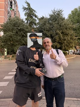 Two men pose for a photo on a Madrid street, one wearing a black balaclava covering his face while holding up a phone, the other in a white shirt smiling and giving a thumbs-up. The image relates to the post's caution about phone theft in Madrid, where the author recounts losing their phone in an Uber after a match and nearly giving up hope of getting it back.
