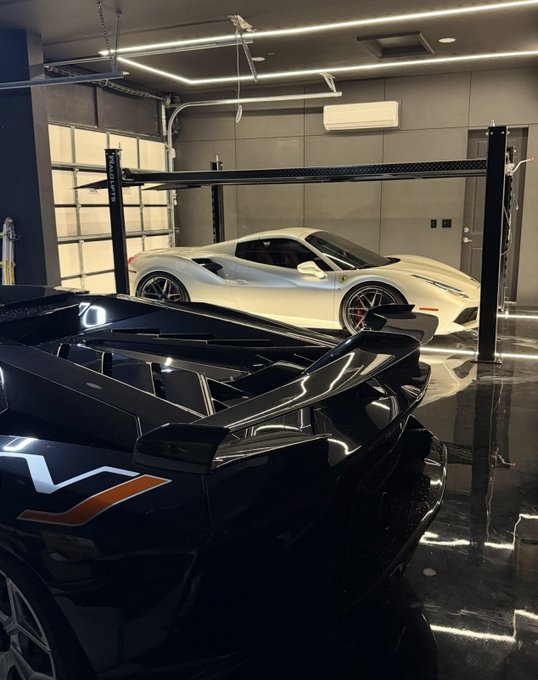 A luxury garage featuring a white Ferrari in the background and a black sports car with a prominent rear wing in the foreground, illuminated by modern ceiling lights.