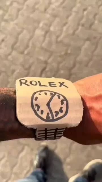 A close-up shows a handmade-looking wristwatch or wristband labeled “ROLEX” with a simple clock drawing on a light square face, worn over a textured outdoor ground.