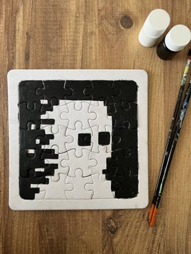 A small completed jigsaw puzzle on a wooden surface, depicting a pixelated black and white face, accompanied by two paintbrushes and small paint bottles.