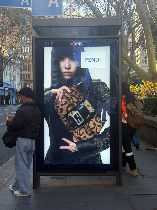 A person wearing a black beanie and light blue jeans stands next to a street-side advertising billboard displaying a FENDI fashion ad featuring a model holding a leopard-print bag, with a "QMS" logo at the top. The accompanying post jokingly claims a "MINA" sighting in Sydney, playfully referencing the striking visual moment of spotting the model in the wild.

Key text visible: "FENDI", "QMS", "A" (on blue signs above the billboard)