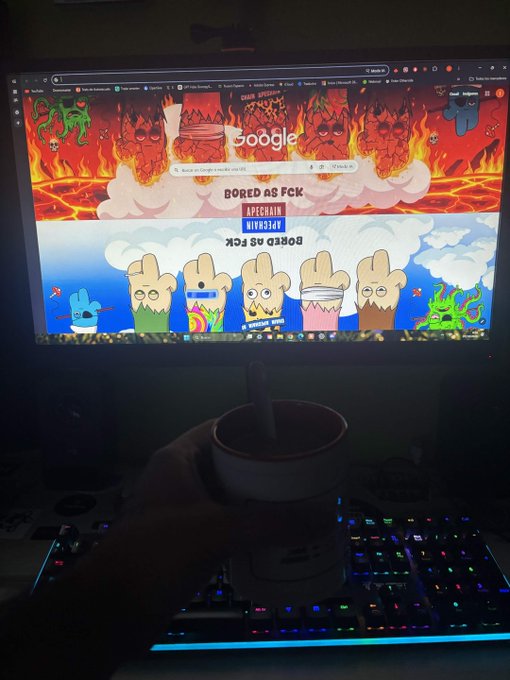A person holding mate (a traditional South American drink) in a gourd in front of a computer displaying a Google homepage with a custom wallpaper featuring pixel-art characters and 'BORED AS FCK' text.