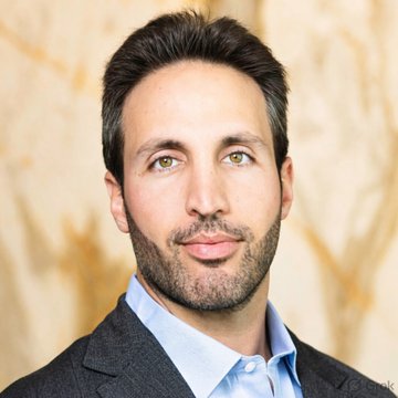 A professional headshot of a man with short dark hair, a trimmed beard, and light-colored eyes, wearing a light blue collared shirt under a dark gray blazer against a neutral beige background—appearing to be Kyle Samani, the Multicoin Capital co-founder referenced in the post about his new board appointment at crypto firm Backpack US.