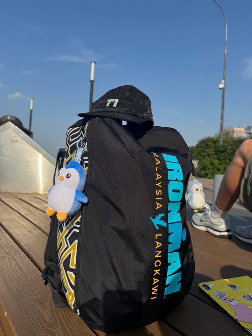 A black "Malaysia Langkawi Ironman" backpack with a cap perched on top and two small penguin plushies attached sits on a wooden deck, accompanied by a caption celebrating the athlete's two Ironman 70.3 finishes and joking that this gear-filled look is how "normies" see them on race days.