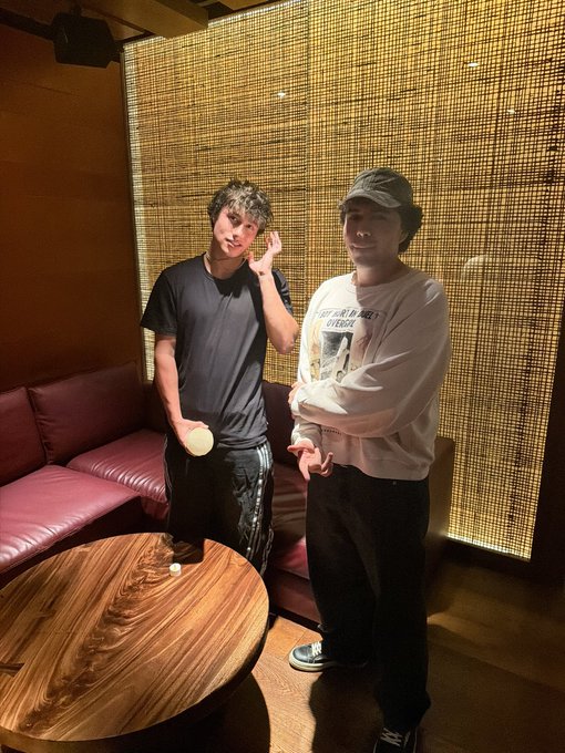 Two young men pose in a private lounge with a bamboo-screened wall and red leather seating, with one wearing a white shirt reading "THE WORLD IS NOT ENOUGH" and the other in a black shirt holding a drink. The caption confirms they've secured a private room, matching the intimate, exclusive setting shown.
